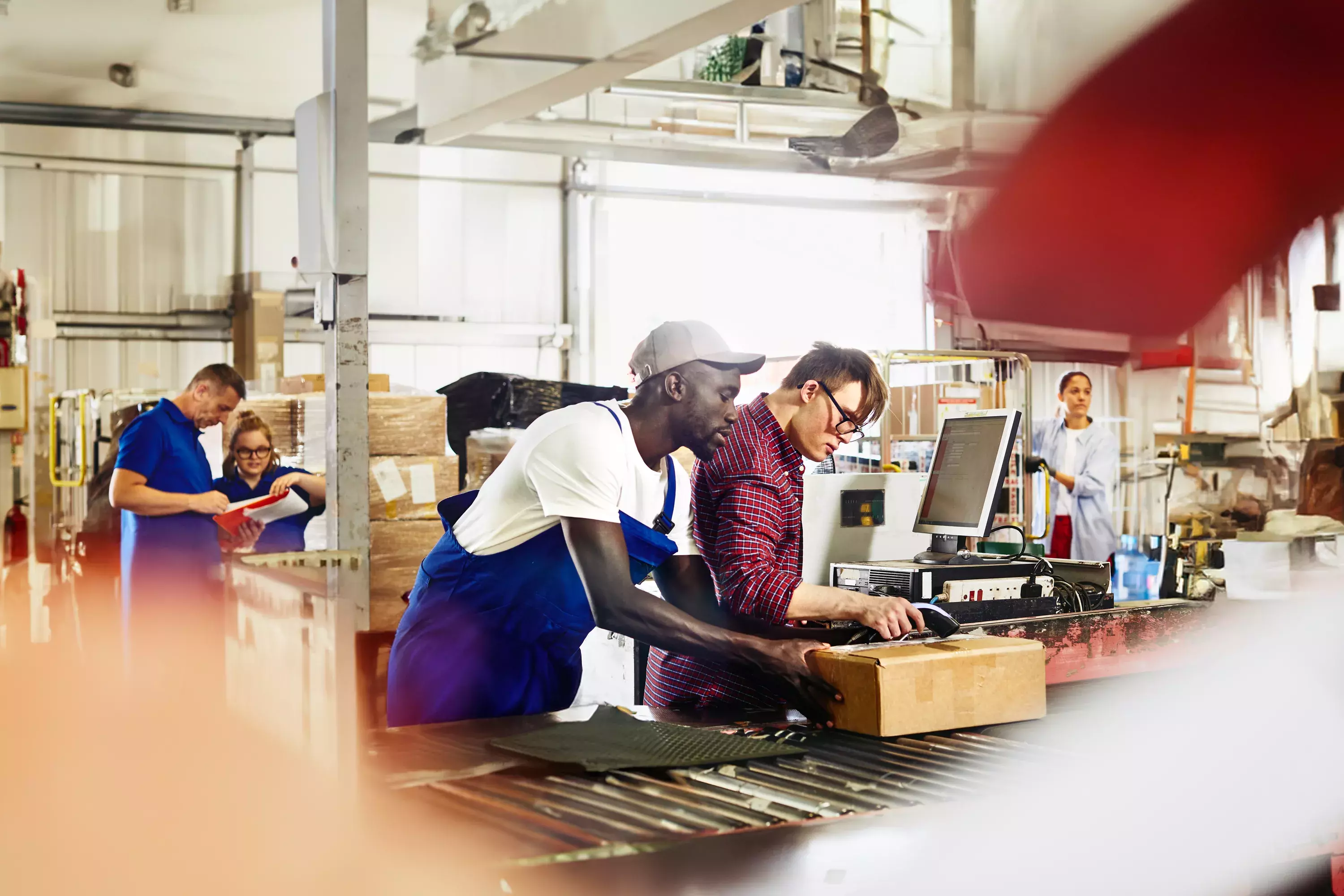 People working together next to a assembly line. male holding a package while other male scans the package. People talking together in the background.
