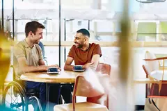 Two men sitting at a table having a conversation. Coffee cups on table.
