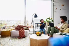 male sitting on couch with his laptop, smiling at a kid holding a notebook.
