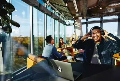 Smiling male with headphones and laptop sitting in a lounge environment.
