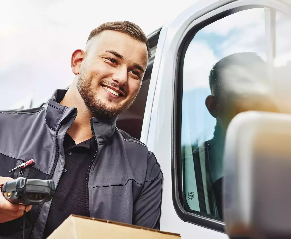 Close up - Smiling male holding a scanner and package standing next to a delivery van.
