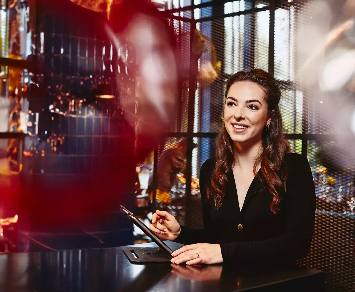 Smiling restaurant hostess (female) looking away, behind the reception desk with tablet.
