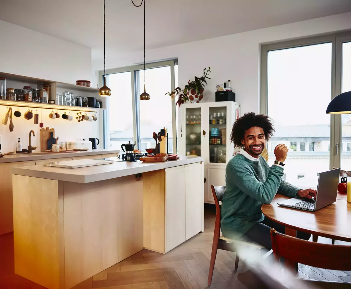 Smiling male sitting at his dining table with his laptop. Kitchen in the background.

