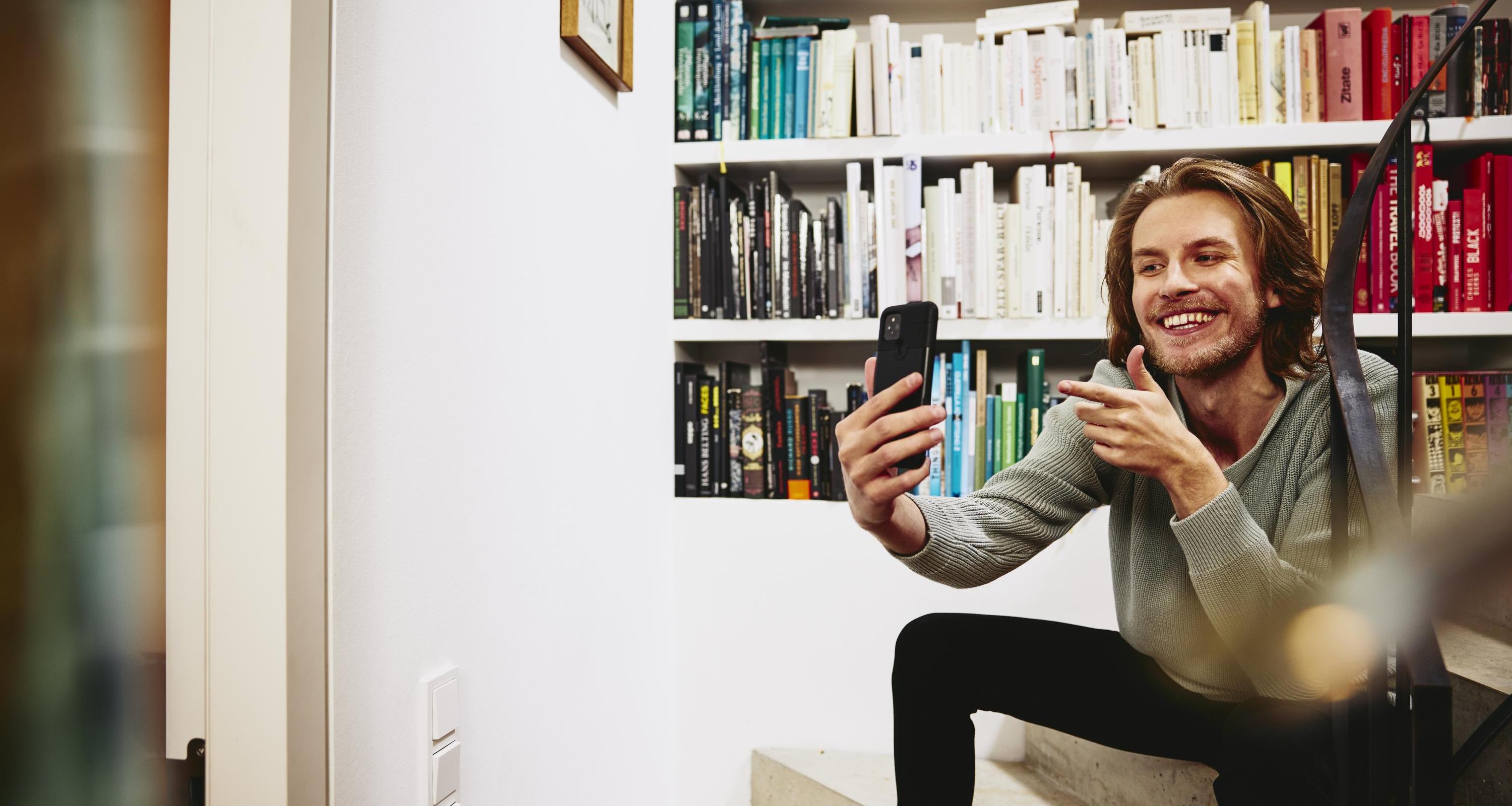 Smiling man sitting on stairs, making a selfie. bookshelf in background