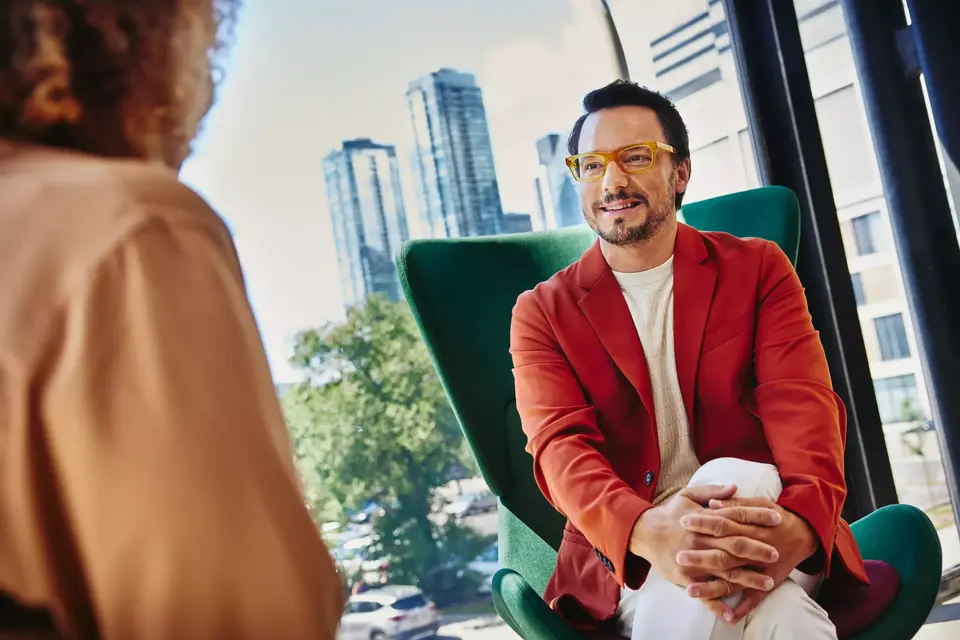 Male with glass and red blazer sitting in a green chair looking at a person