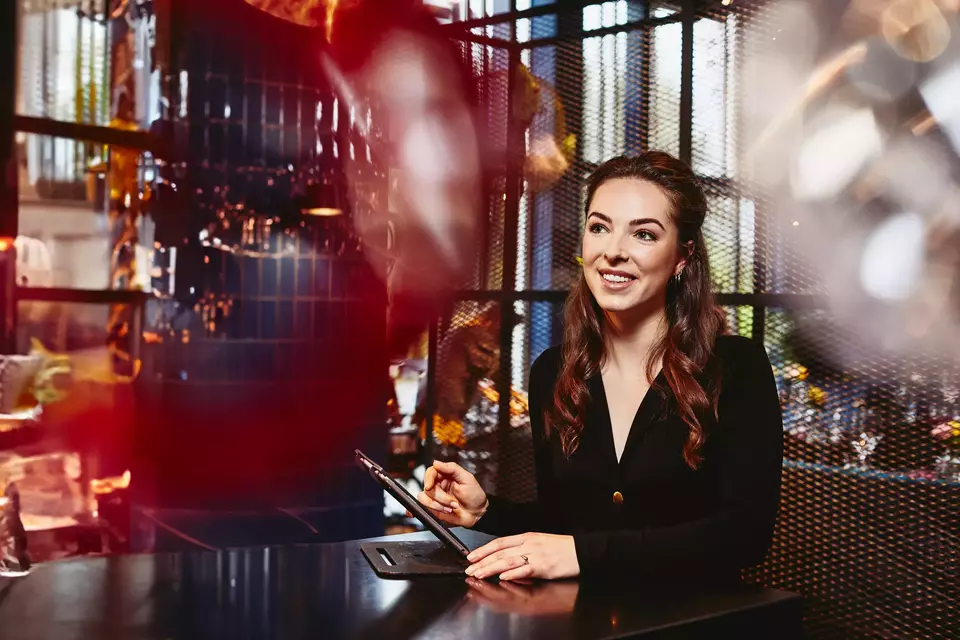 Smiling restaurant hostess (female) looking away, behind the reception desk with tablet.
