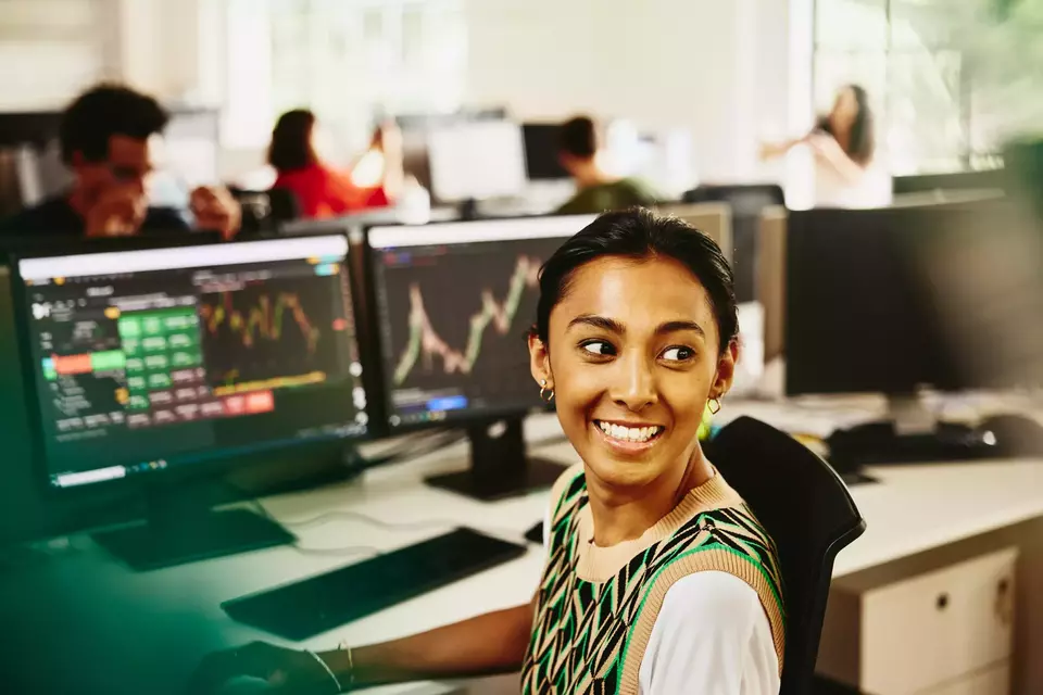 Smiling female looking away from computer screens displaying financial information.
